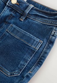 Jeans de mezclilla con un acabado azul, que presentan un bolsillo frontal, costuras visibles y un botón metálico. La tela tiene una textura suave.