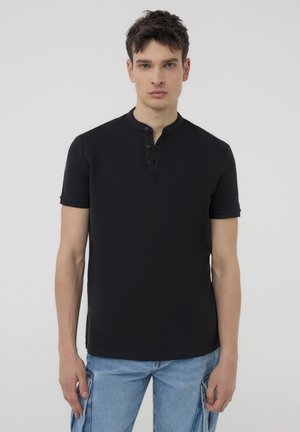 Polo shirt nero a maniche corte con colletto arrotondato e patta con tre bottoni, realizzato in tessuto liscio. Indossato con pantaloni cargo blu chiaro.