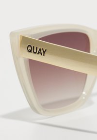 Sončna očala s prozornimi bež okvirji, gradientnimi roza lečami in zlatimi nosilci, na katerih je v črnih črkah vgravirano "QUAY".
