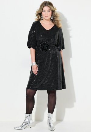 Schwarzes Paillettenkleid mit V-Ausschnitt und kurzen Ärmeln, ausgestattet mit einem schwarzen Blumen-Gürtel. Stylisch kombiniert mit silbernen Ankle Boots und Netzstrumpfhosen.