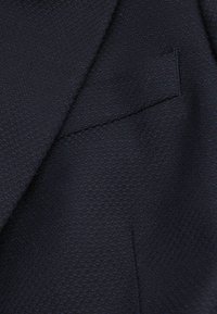 Close-up af mørkt tekstureret stof på en skræddersyet jakkesætjakke med en hakket revers og en brystlomme.