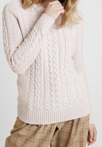 Rosa kabelstickad tröja med ribbade ärmslut och nederkant, texturerade mönster och en avslappnad passform, i kombination med bruna rutiga byxor.