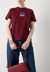 Bordeaux bomull t-skjorte med en firkantet lilla logo. Kombinert med vide blå jeans og en rød lærveske holdt av en sølvdetaljert hånd.
