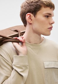 Béžová mikina s dlhými rukávmi a náprsným vreckom. Model nesie hnedú tašku s koženými remienkami, ktorá ukazuje hladkú textúru a jednoduchý dizajn.