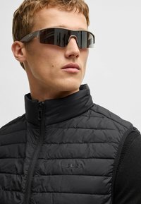Schwarze Steppweste mit hohem Kragen, mit gestepptem Textil und Reißverschluss. Eine Sonnenbrille mit braunem Gestell und dunklen Gläsern rundet den Look ab.