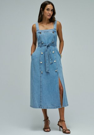 Mujer que lleva un vestido midi de denim azul sin mangas con botones delanteros, cinturón en la cintura, abertura lateral y sandalias marrones de tacón alto sobre un fondo gris liso.