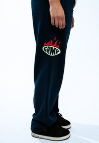 Granatowe dresy o luźnym kroju z dużym zielono-czerwonym logo płomienia i napisem "GRMC" na lewym udzie. Czarne sneakersy.