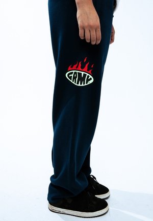 Sudadera azul marino con un corte relajado que presenta un gran logotipo de llamas en verde y rojo con el texto "GRMC" en el muslo izquierdo. Zapatillas negras.
