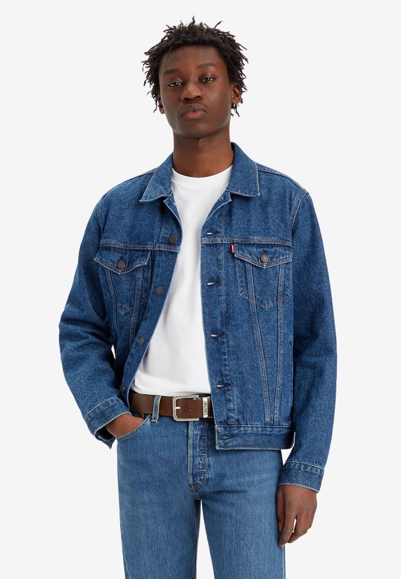 Veste en denim bleu foncé au design classique, avec fermeture à boutons sur le devant, deux poches poitrine et des coutures contrastées. Associée à une chemise blanche.