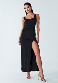 Črna maxi obleka s kvadratnim izrezom, nabranim pasom in razporkom do stegna. Kombinirana z zlato usnjenimi sandali z visokimi petami. Gladek tekstura.