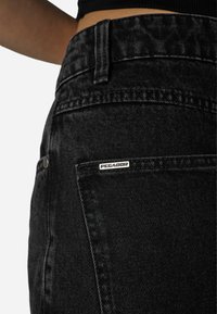 Crne traperice od denima s teksturiranim završetkom, s jednim stražnjim džepom i metalnom oznakom s natpisom "PEGADOR." Krupni plan detalja tkanine od denima.