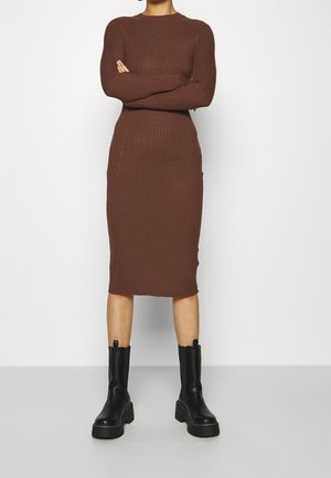 Persoon die een bruine midi-jurk met geribbelde stof en lange mouwen draagt, samen met zwarte grove enkellaarsjes, staand met gekruiste armen.