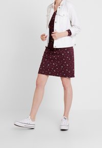 Burgunderfarbendes Blumenkleid mit kurzen Ärmeln, kombiniert mit einer weißen Jeansjacke und weißen Sneakers. Weicher Stoff, knielanger Schnitt.