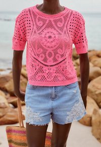 Jasnoróżowa, szydełkowana bluzka z krótkim rękawem z misternymi wzorami. Połączona z dżinsowymi szortami z kwiatowymi haftami.