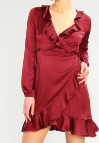 Robe portefeuille en satin bordeaux avec manches longues, décolleté froncé et ourlet évasé. Présente une taille cintrée et un détail de lien sur le côté.
