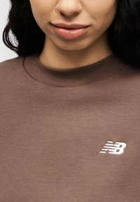 Καφέ φούτερ με ριμπ λαιμόκοψη και λευκό κεντημένο λογότυπο στο στήθος. Απαλή υφή, καθημερινός σχεδιασμός.