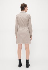 Abito beige chiaro con vita cinturata, maniche lunghe e colletto. Tessuto di cotone liscio con taglio dritto, che arriva a metà coscia. Stivali neri alla caviglia.