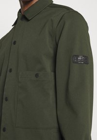 Close-up van een donkergroene jas met zwarte drukknopen, klepzak, puntige kraag en zwarte Barbour International patch op de bovenmouw.