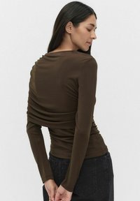 Ruskea pitkähihaine paita, jossa on kerätty kangasosa selässä. Sujuva pinta ja istuva muotoilu, yhdistettynä tummiin farkkuihin.