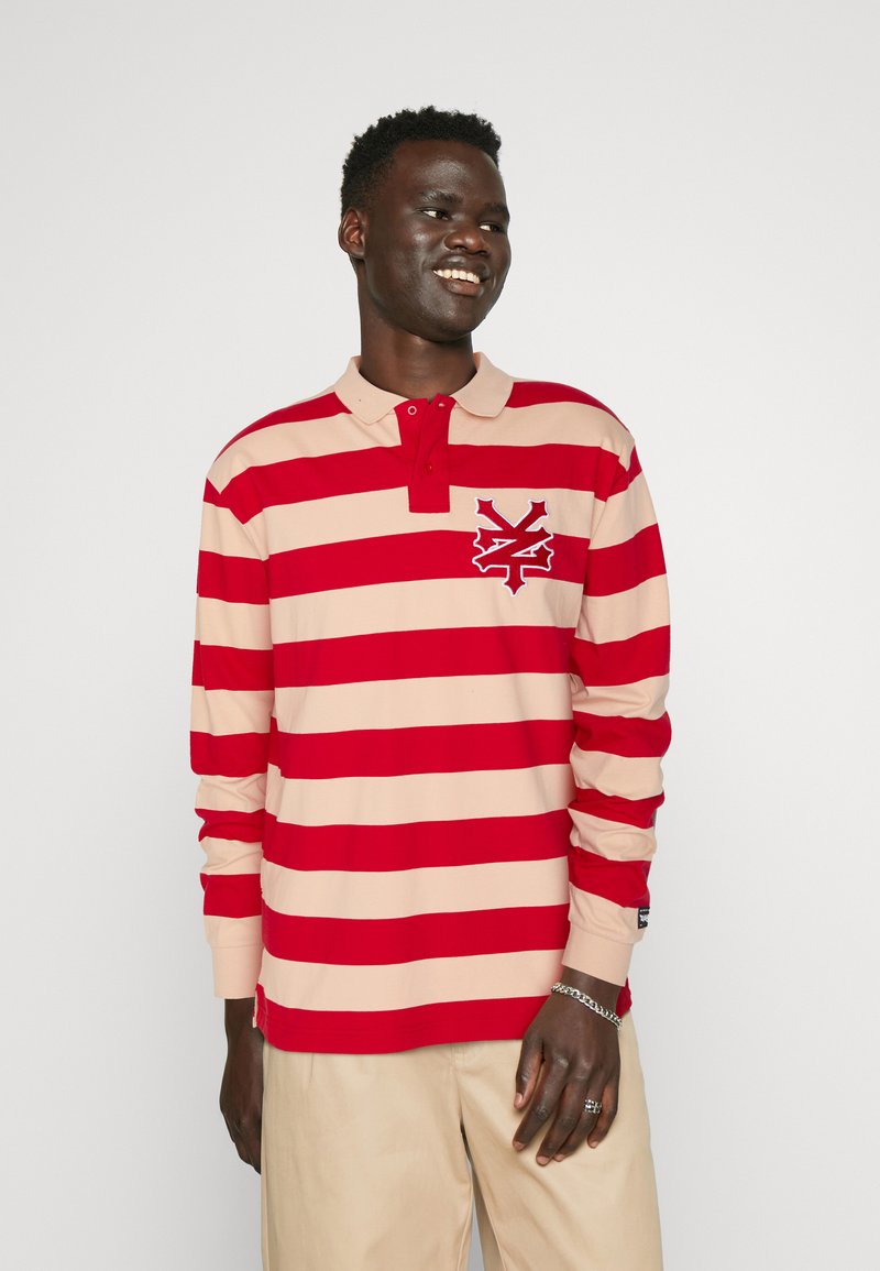 Long-sleeve polo shirt in beige with wide red stripes, featuring a collar, three-button placket, and a logo on the left chest.