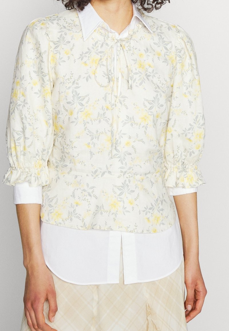 Vit blommig blus med gula och grå mönster, puffärmar, knytning i halsen och en vit skjortkrage som är lager på lager under.