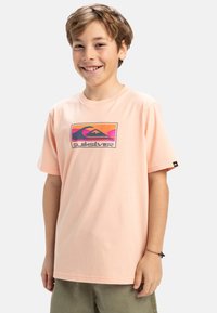 Ljus persikofärgad bomullströja med korta ärmar och rund halsringning, med en färgglad Quiksilver-logotyp på framsidan.