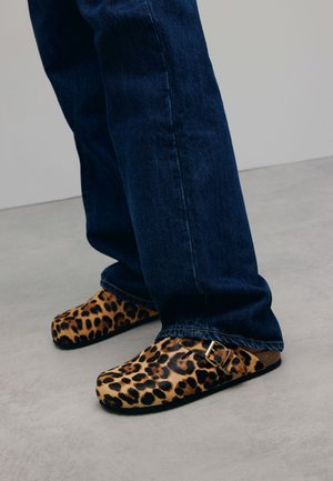 Leopardprint klompen van een gestructureerde stof, voorzien van een gespdetail, gecombineerd met donkerblauwe spijkerbroeken.