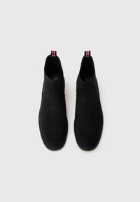 Botines de ante negro hasta el tobillo con punta redonda. Cuentan con un lazo decorativo de rayas rojas, blancas y azules en la parte trasera y una suela plana de goma.