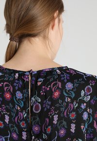 Svart blommig blus med livfull brodyr i lila, röd och blå. Nyckelhålsdetalj i ryggen med rynkad halsringning och mjuk textur.