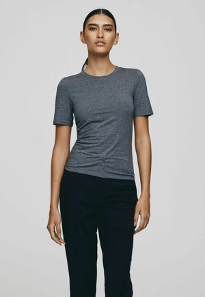 Woman standing wearing a fitted short-sleeve gray t-shirt and black pants against a plain light gray background.