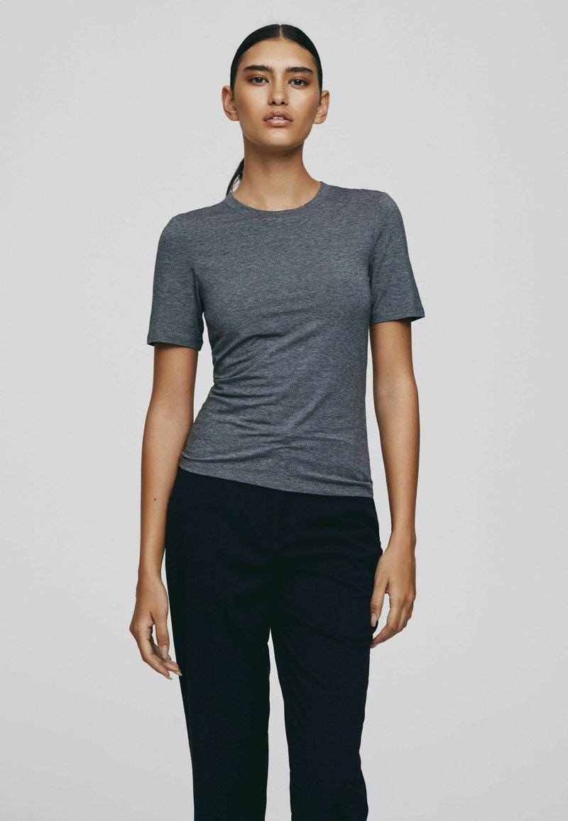 Naine seisab, kandes kehale istuvat lühikeste varrukatega halli t-särki ja musti pükse, taustaks lihtne heledat halli värvi taust.