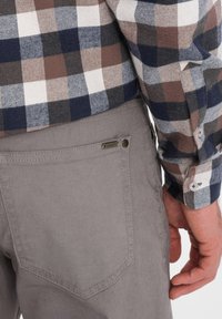 Un pantalon en coton gris présente une poche à l'arrière avec des coutures et une petite étiquette en métal. Une chemise à carreaux marron et marine est superposée.