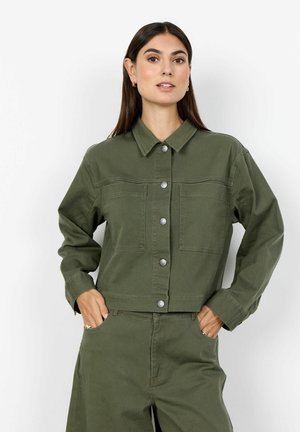 Vrouw met lang donker haar, dragende een dichtgeknoopte olijfgroene jas en bijpassende broek, met handen in de zakken tegen een witte achtergrond.
