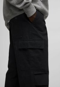 Czarne spodnie cargo wykonane z wytrzymałego materiału, z wieloma dużymi kieszeniami i gładką fakturą, stylizowane na dopasowany krój.