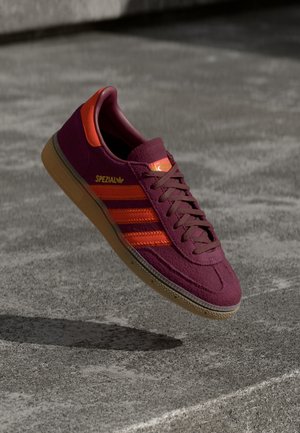 Bordowe buty sportowe Adidas Spezial z czerwonymi paskami i gumową podeszwą unoszące się nad betonową nawierzchnią.