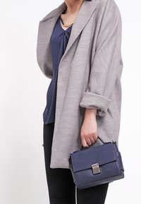 Grå cardigan med uppkavlade ärmar över en mörkblå topp, håller en liten marinblå handväska med texturerad finish och metallspänne.