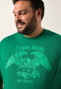 Muž s vousy se usmívá, má na sobě zelené tričko s grafikou orla a textem "Denim Brand EST. 2007 New York City."