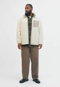 Giacca esterna in pile crema con zip frontale, forma oversize, due grandi tasche e una tasca patch grigia con dettaglio del logo.