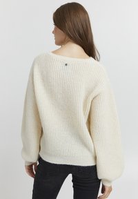 Pull en tricot couleur crème avec une coupe ample, épaules tombantes, texture côtelée et un petit bouton décoratif au dos du cou.