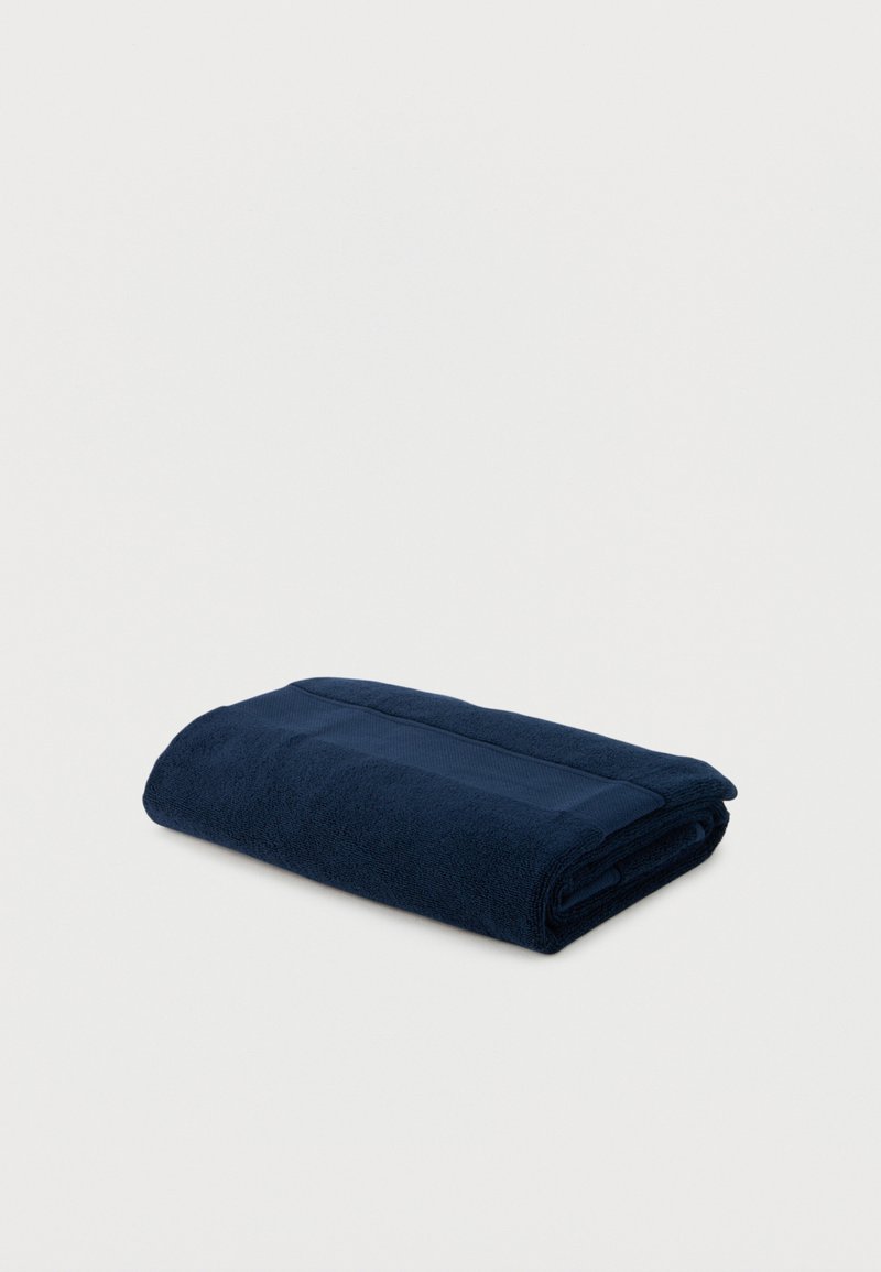 Opgevouwen donkerblauwe katoenen handdoek plat liggend op een lichte ondergrond met een gestructureerde horizontale streep nabij de rand.