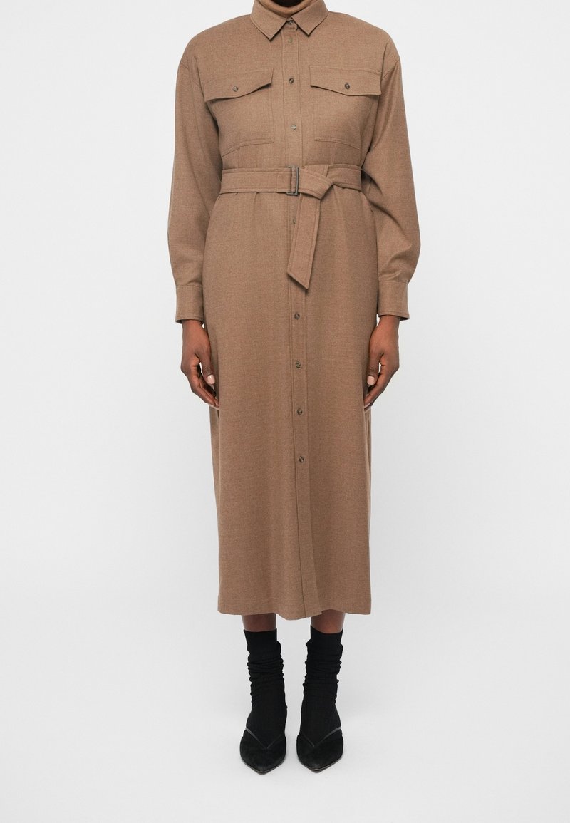 Robe midi marron à manches longues avec ceinture, boutons sur le devant, poches à rabat sur la poitrine, portée avec des chaussettes noires et des talons à bout pointu.