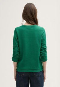 Zeleni sweatshirt s teksturama, okruglim izrezom i dugim rukavima, s rebrastim rubom i manžetama. Nošen uz tamne traperice.