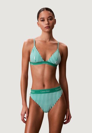 Kobieta ubrana w zielono-biały pionowo prążkowany bikini marki Calvin Klein z trójkątnym stanikiem i majtkami z wysokimi wycięciami, stojąca na jednolitym tle.