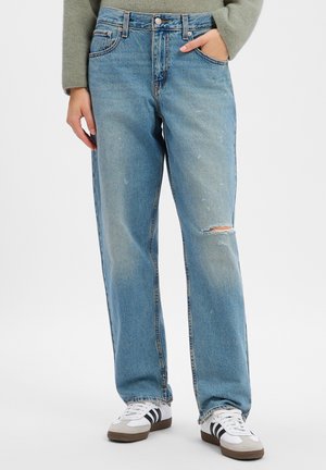 Personne portant un jean droit déchiré bleu clair avec les mains dans les poches et des baskets blanches avec des rayures noires.