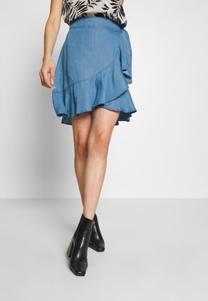 Femme portant une jupe portefeuille bleue à volants associée à des bottines à talons noires, debout devant un fond clair uni.