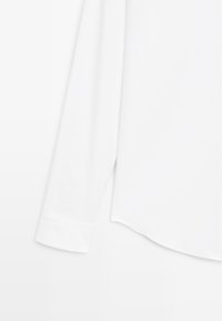 Camicia bianca a maniche lunghe con texture liscia, orlo stondato e polsini con bottone. Il tessuto sembra leggero e traspirante.