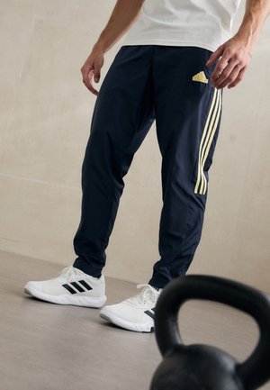 Persona con pantalones deportivos Adidas azul marino con franjas amarillas y zapatillas Adidas blancas, de pie junto a una pesa rusa en el suelo.