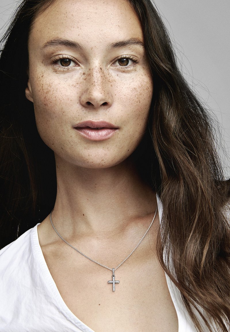 Collana a croce argentata su una delicata catena indossata sopra una camicia bianca. Il ciondolo presenta dettagli sottili e riflette la luce in modo morbido.