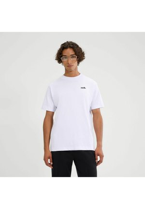 Junger Mann mit lockigem Haar und Brille, der ein schlichtes weißes T-Shirt mit kleinem schwarzem Logo auf der Brust und schwarze Hosen vor weißem Hintergrund trägt.