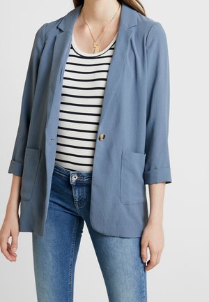 Kobieta w jasnoniebieskiej marynarce na czarno-białym paskowanym topie oraz niebieskich dżinsach, widoczna od szyi do bioder, neutralne tło.